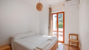 Een slaapkamer met een bed, een houten nachtkastje, een rieten stoel, een raam met gordijnen en een airconditioner.