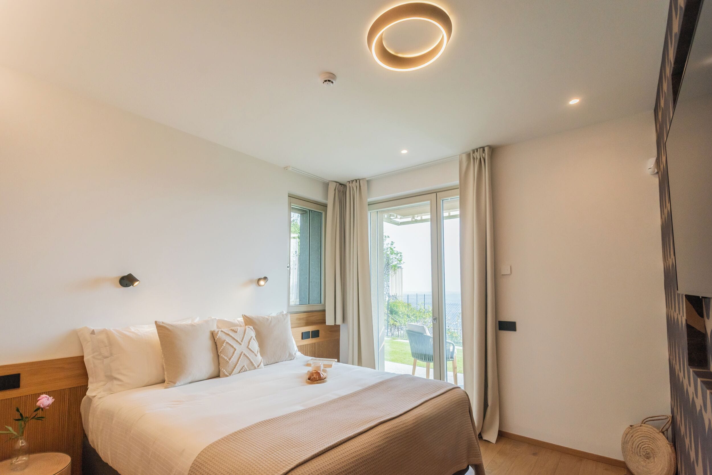 A modern hotel room with a large bed, a small round table, a vase with flowers, and a view of the outdoors through a sliding glass door.