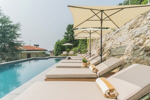 Zona de piscina con sillones de descanso y sombrillas, una pared de piedra y vista a un edificio con tejado de azulejos.