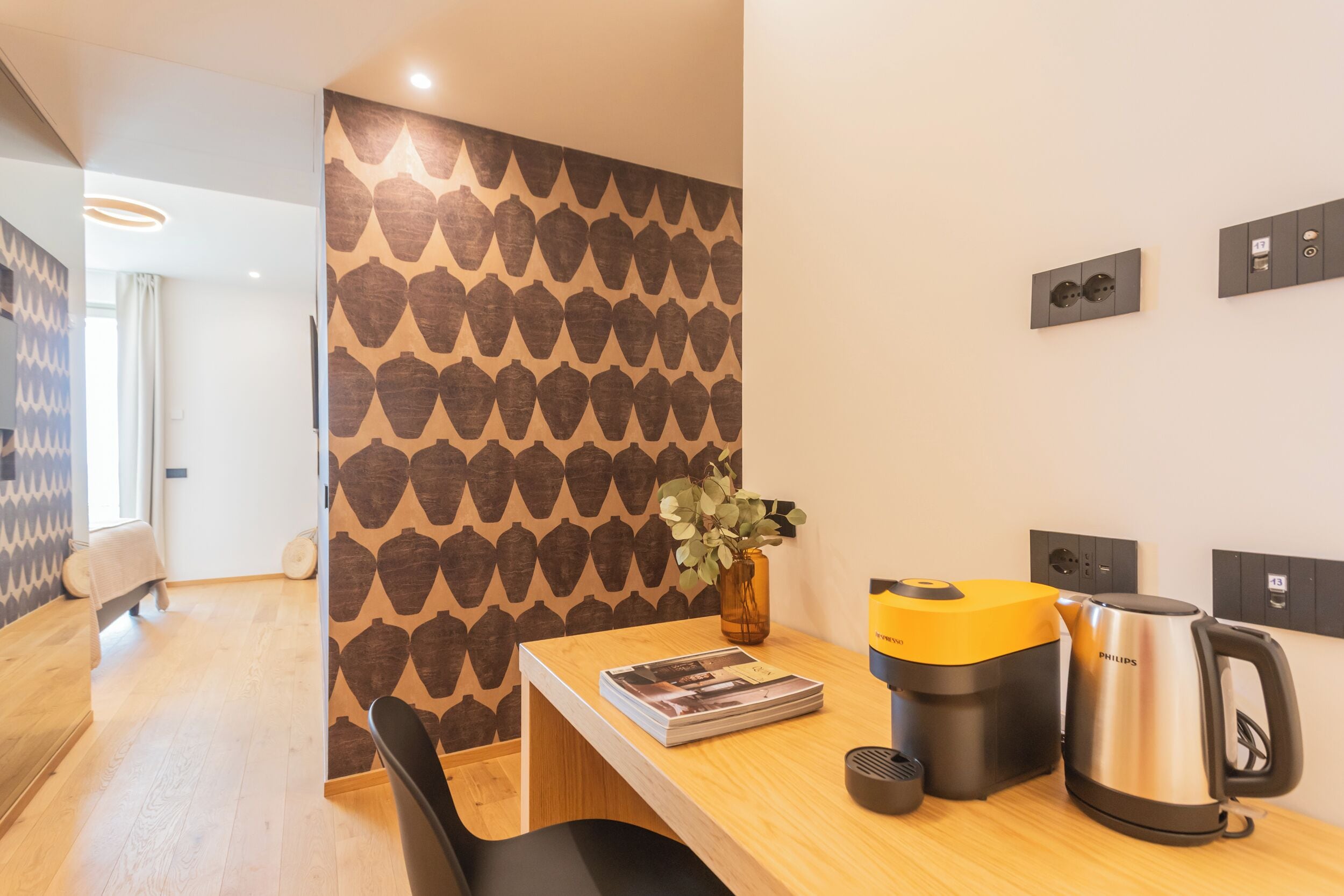 Ein modernes Hotelzimmer mit einem Holztisch, einer Zeitschrift, einer gelben Kaffeemaschine, einem schwarzen Stuhl und einer Wand mit geometrischem Muster.