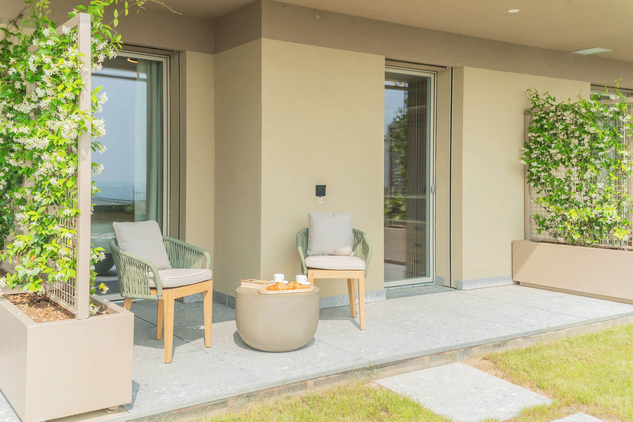Un espace extérieur moderne aménagé avec deux chaises et une table ronde, entouré de plantes en pot et doté d’une porte vitrée donnant sur un balcon.