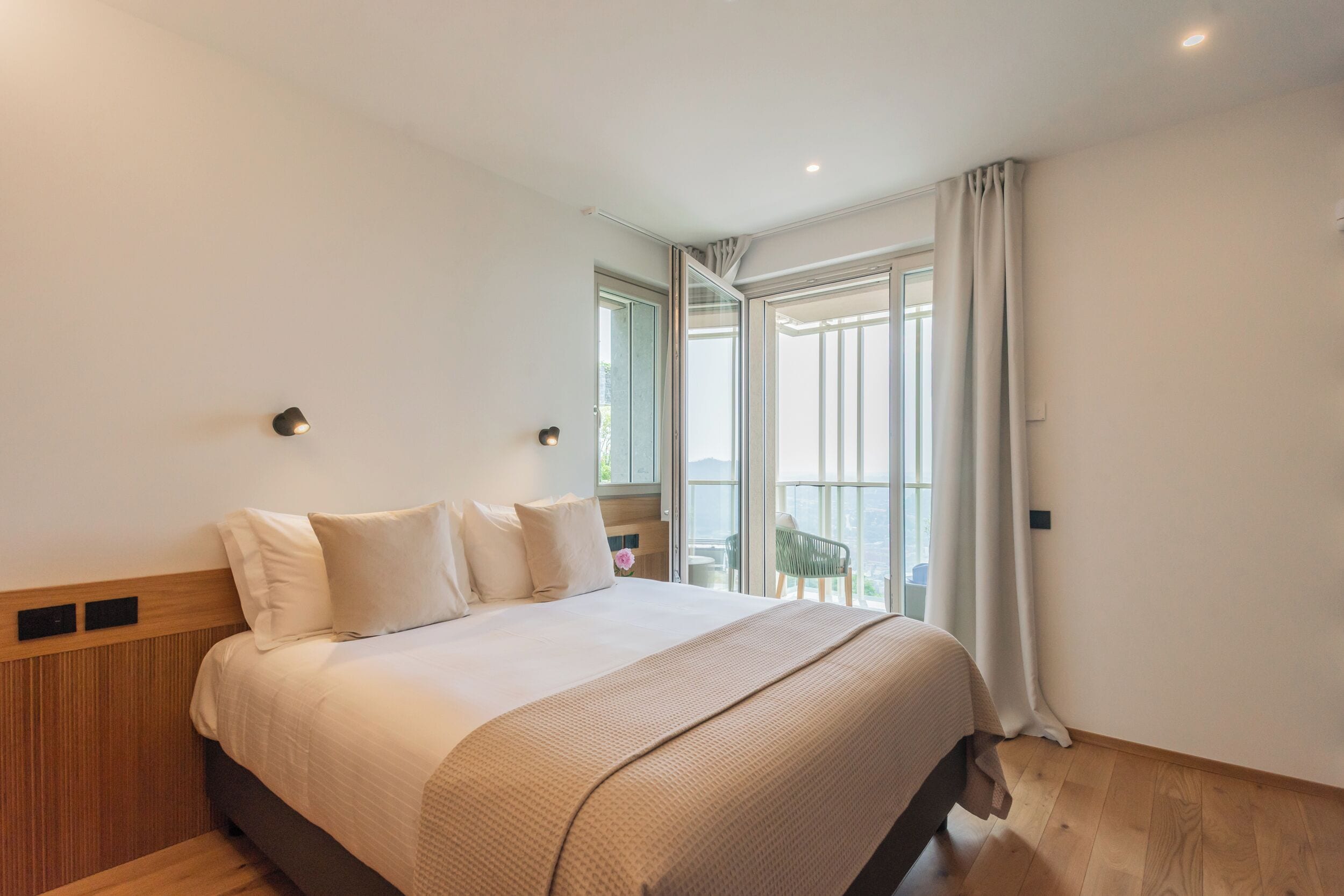 Un dormitorio con una cama grande, un cabecero de madera, una mesita de noche y vistas al exterior a través de un ventanal.