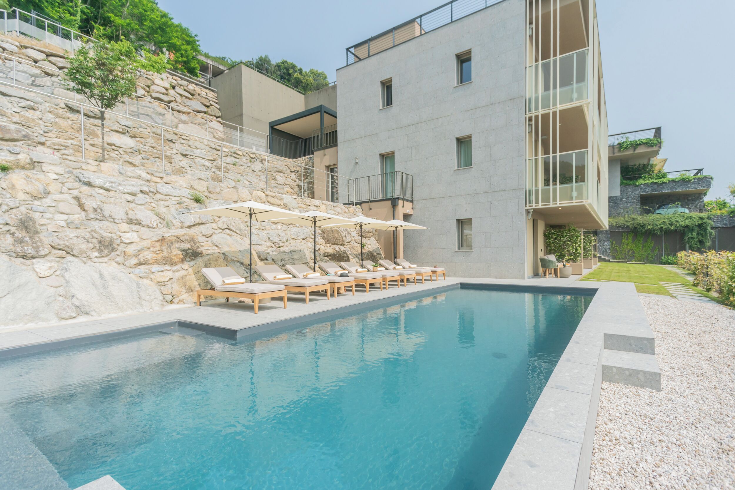 Ein moderner Poolbereich mit Liegestühlen und Sonnenschirmen, umgeben von Steinmauern und einem mehrstöckigen Gebäude mit Balkonen.