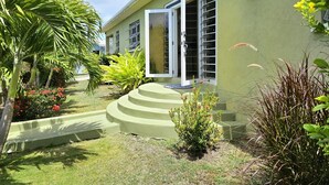 Un edificio con fachada verde, una escalera que lleva a una puerta y un pequeño jardín con diversas plantas y una palmera.