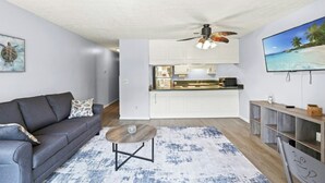 A modern living room with a grey sofa, a coffee table, and a kitchen area with white cabinets and a TV displaying a beach scene. - Shipwatch Pointe II-K103 1BR Condo in Shore Dr (Myrtle Beach)