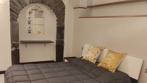 Une chambre avec une arche en pierre, un lit recouvert d’une couverture grise, deux oreillers ornés de motifs de feuilles, une table de chevet et une commode.