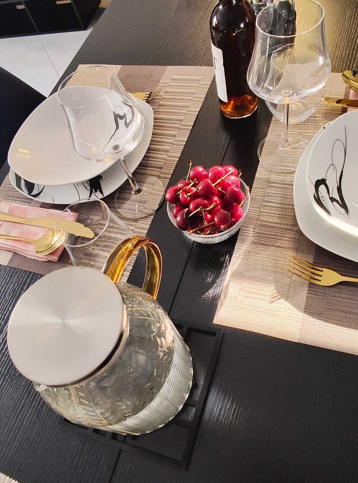 Una mesa de comedor dispuesta con platos, cubiertos, una jarra de cristal, un bol de cerezas y una botella de vino.