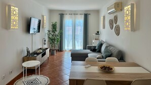 A modern living room with a sofa, dining table, and a TV. There is a potted plant and a small table with decorative items. - Morgado Group - Quinta do Morgado - Morgado Privilege (Tavira)