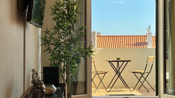 A living room with a television, a small table with a remote control, a decorative plant, and a view of a tiled patio outside. - Morgado Group - Quinta do Morgado - Morgado Privilege (Tavira)