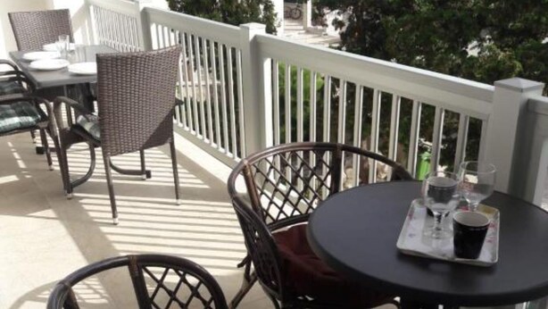 A balcony with wicker chairs and a table, a white railing, and a view of trees and a bicycle in the background.