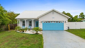 A single-story house with a blue garage door, a small porch with two chairs, and a number 132 on the front. - Beach House - Baby Blue Heron by PHG (Panama City Beach)