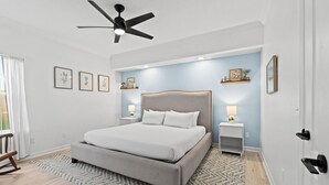 A bedroom with a bed, bedside tables, a ceiling fan, and wall-mounted shelves. - Beach House - Baby Blue Heron by PHG (Panama City Beach)