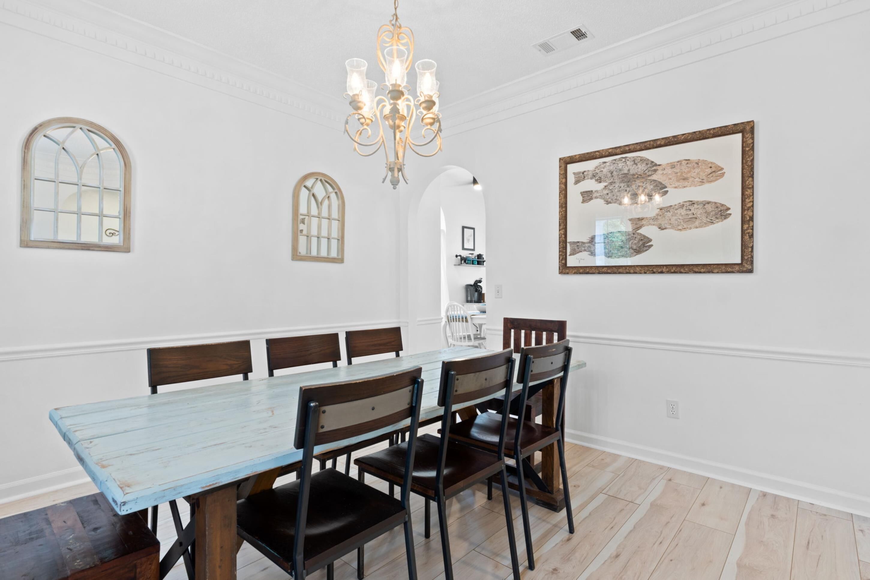 Uma sala de jantar com uma mesa comprida, seis cadeiras, um candelabro e uma obra de arte emoldurada na parede.