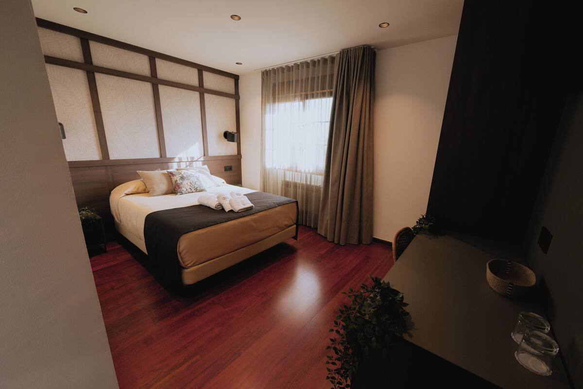 Une chambre d’hôtel moderne avec un grand lit, un plancher en bois, une table de chevet avec une lampe et une grande fenêtre avec des rideaux.