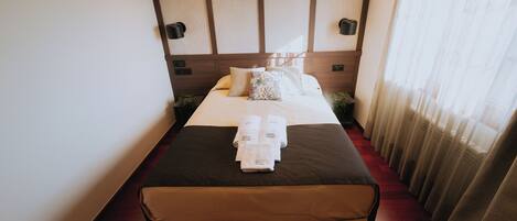 Ein Hotelzimmer mit einem großen Bett, einem Blattmuster-Kopfbrett, zwei Wandlampen und einem Fenster mit durchsichtigen Vorhängen.