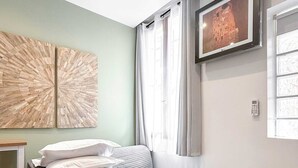 Un dormitorio moderno con un sofá cama gris, dos obras de arte en la pared, un televisor y una ventana con cortinas.