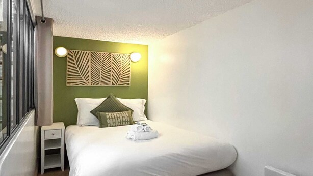 Un dormitorio pequeño y ordenado con cama, mesita de noche, luz de pared y ventana.