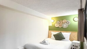 Una cama bien hecha con sábanas blancas y una toalla doblada, una mesita de noche con cajón, y una lámpara fijada a la pared.