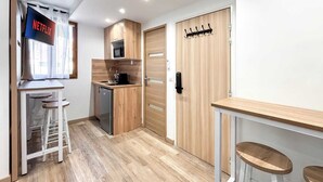 Eine kompakte Küche mit Mikrowelle, Spüle und kleinem Kühlschrank. Eine Holztür mit Kleiderhaken, und ein an der Wand befestigtes Fernsehgerät, das 'NETFLIX' anzeigt.