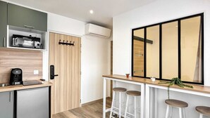Eine kleine Küchenzeile mit Mikrowelle, Kaffeemaschine und kleinem Kühlschrank. Drei weiße Barhocker stehen vor einer Holzausstattung, über der sich ein großer Spiegel befindet.