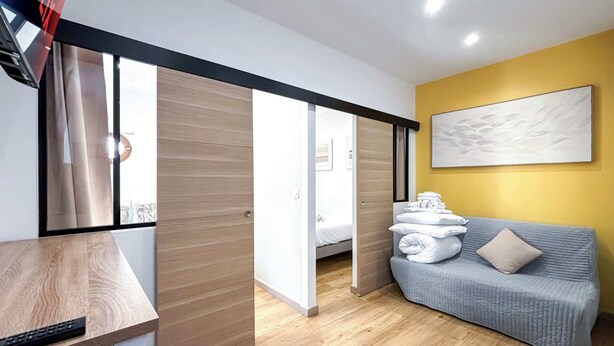 Quarto de hotel moderno com piso de madeira, um sofá cinza, uma cama com roupa branca e um quadro na parede.