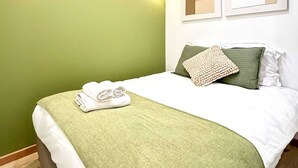 Un lit bien fait, avec un plaid vert et des oreillers, une serviette pliée, et deux œuvres d’art encadrées accrochées au mur.