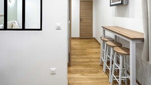 Een hal met een houten vloer, een witte muur met een afbeelding in een lijst, een kleine tafel met krukjes en een deur aan het einde.