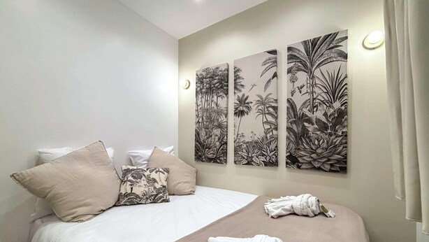 Ein ordentlich bezogenes Bett mit einer beige farbenen Tagesdecke und Kissen, einem Wandbild im tropischen Stil und einem Handtuch auf dem Bett.