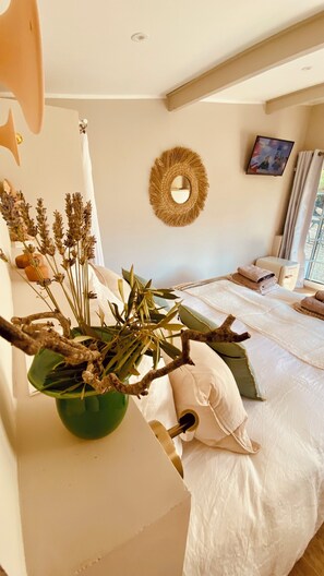 A bedroom with a bed, a green vase with dried plants, a wall-mounted mirror, and a window with curtains.