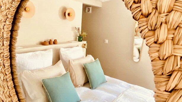 Un espejo circular rodeado por un marco tejido, que refleja una cama con almohadas blancas y azul turquesa, una mesita de noche con una planta, y una lámpara fijada a la pared.