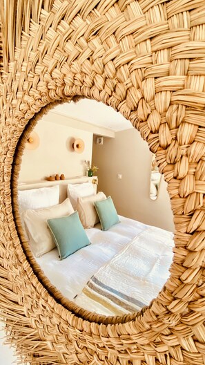 A circular mirror surrounded by a woven frame, reflecting a bed with white and teal pillows, a bedside table with a plant, and a wall-mounted light fixture.