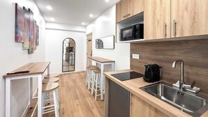 A modern kitchen with wooden cabinets, a stainless steel sink, and a microwave. There are stools and a small table, and the floor is wooden.
