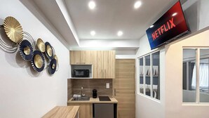 Kompakti keittiö, jossa on mikroaaltouuni, pesuallas ja pieni jääkaappi. Tasomaisen näytön omaava televisio on asennettu työtason yläpuolelle, ja sen näytöllä näkyy Netflixin logo.