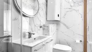 A modern bathroom with a glass shower enclosure, a round mirror, a white sink, a toilet, and a marble backsplash. - Hoche C 105 - Studio Apartment, Sleeps 2 (Paris)