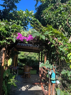 A wooden pergola with a green table and chairs underneath, surrounded by lush greenery and a clear blue sky.
