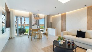 A modern living room with a sofa, a dining table with chairs, and a view of the sea through large windows.