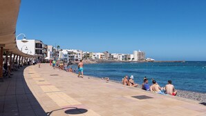 A seaside promenade with people relaxing by the water, buildings in the background, and a clear blue sky.