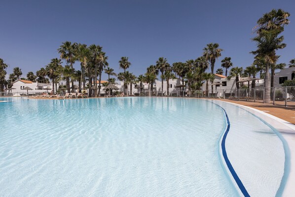 Une piscine aux eaux cristallines, à bords incurvés, entourée de palmiers et de bâtiments blancs aux toits rouges.