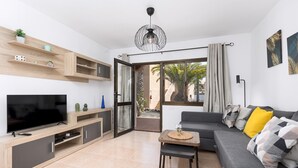 Een moderne woonkamer met een flatscreen televisie, houten planken, een grijze bank met kussens, een bijzettafeltje en een schuifdeur van glas naar een buitenruimte.