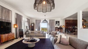 A spacious living room with a large sofa set, a coffee table, and a television. There is a dining area with a wooden cabinet and a lamp. The room has a large window with curtains and a view of the outdoors.
