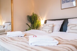 Una cama bien tendida con sábanas y almohadas blancas, una planta en la mesita de noche y una lámpara de pared.