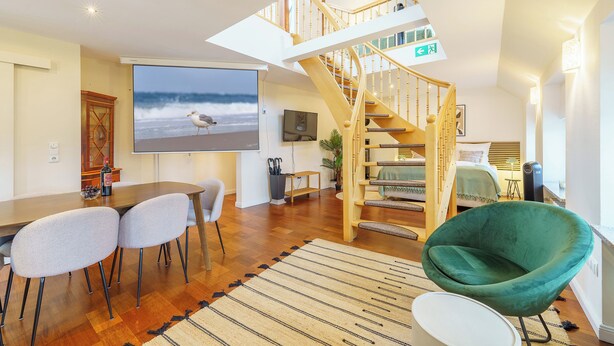 A spacious living area with a wooden staircase, a dining table with chairs, a large TV screen displaying a beach scene, and a green armchair. - Residenz am Hostentor (Lübeck)