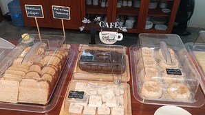 Una vetrina di panetteria con diversi prodotti da forno in contenitori trasparenti, con etichette che riportano nomi come “Pão de Queijo” e “Pão de Queijo de Papel”.