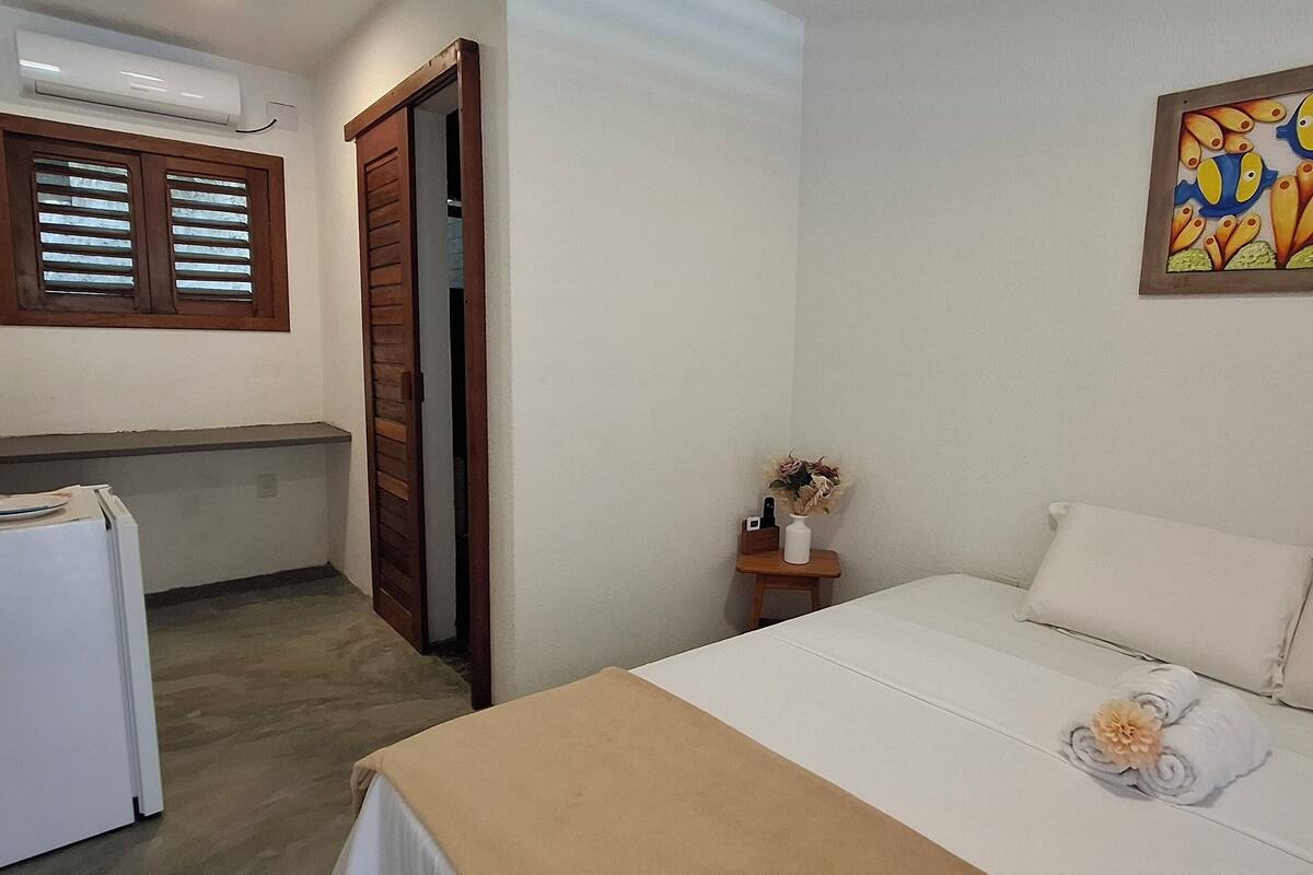 A hotel room with a bed, a small table with a vase, a refrigerator, a window with blinds, and a wall-mounted air conditioner.
