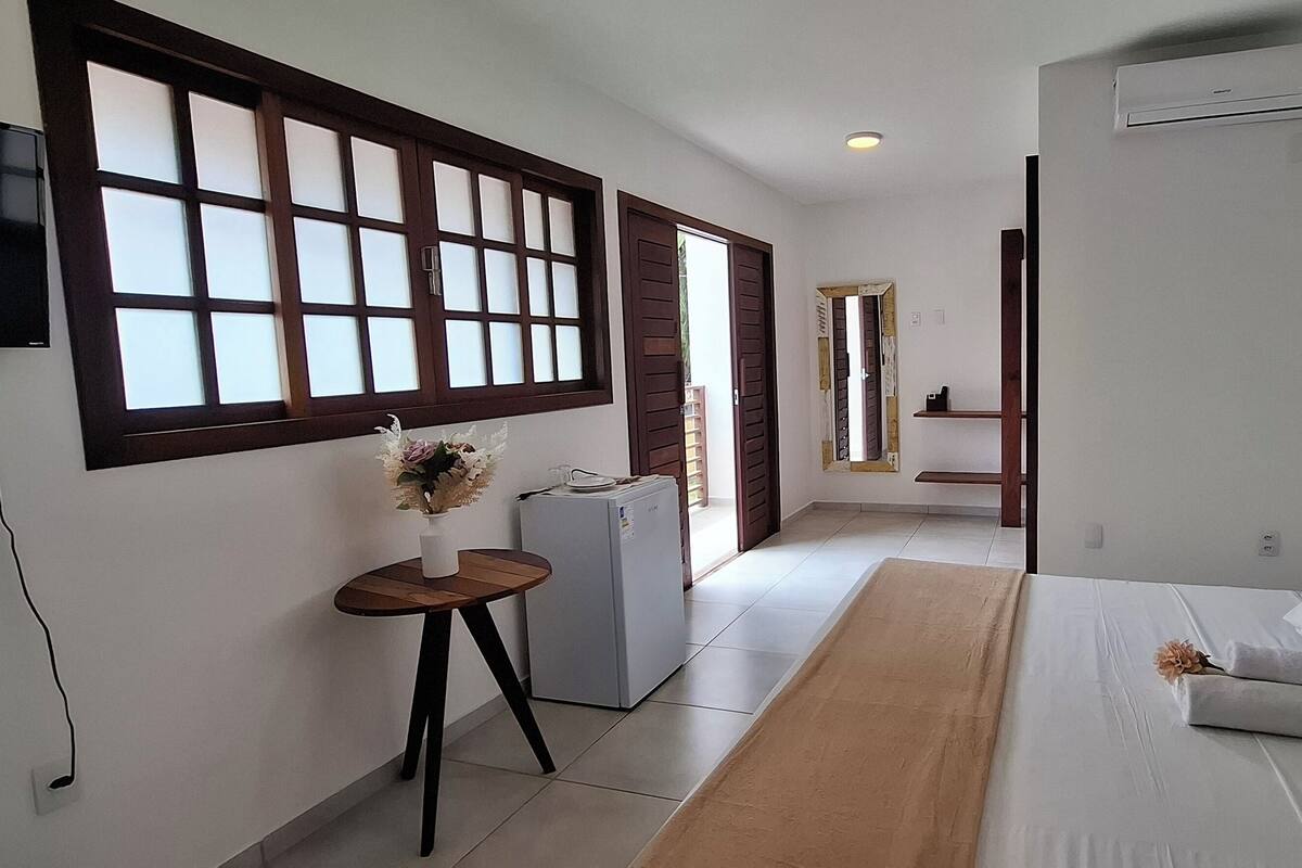 A modern hotel room with a large bed, a small table with a vase, a refrigerator, and a TV mounted on the wall.