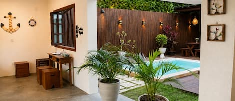 A room with a wooden desk, a potted plant, a window with blinds, and a view of a pool area.