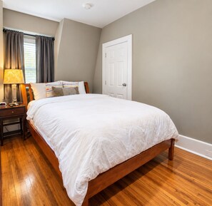 A bedroom with a wooden bed, a nightstand with a lamp, a window with curtains, and a door.