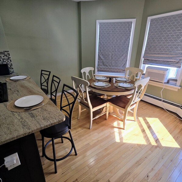 A dining area with a wooden table and chairs, a kitchen counter with a plate and utensils, and a window with a patterned blind.