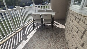 A covered patio area with a table and chairs, a white railing, and a view of the street and houses in the background.