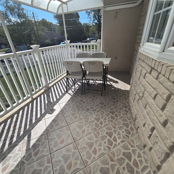 A covered patio area with a table and chairs, a white railing, and a view of the street and houses in the background.